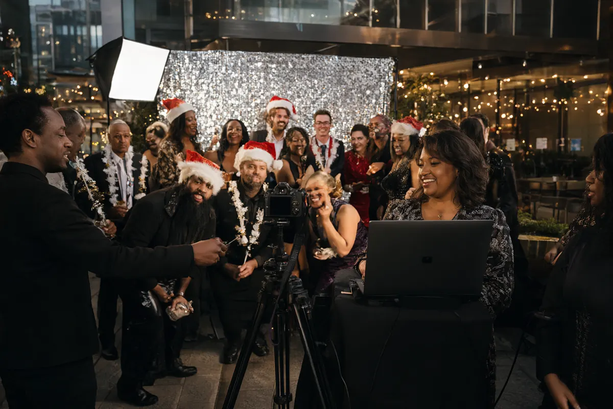 An open-air photo booth with a silver sequin backdrop at a corporate holiday party, an attendant guiding colleagues
