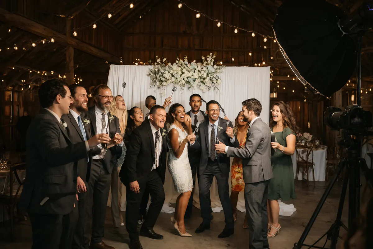 Wedding guests laughing with props in front of a white floral photo booth backdrop in a rustic barn venue