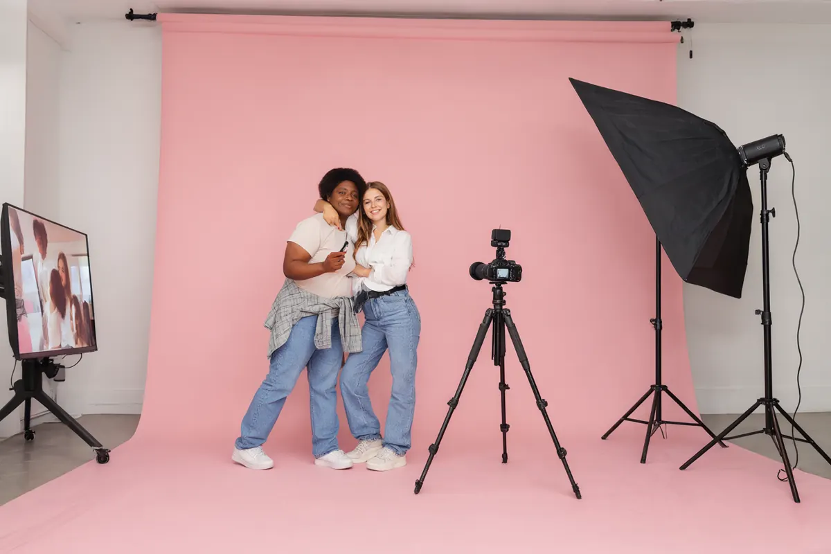 Two friends posing in a minimalist self-portrait studio with a pink backdrop, holding a shutter remote, live view on a monitor
