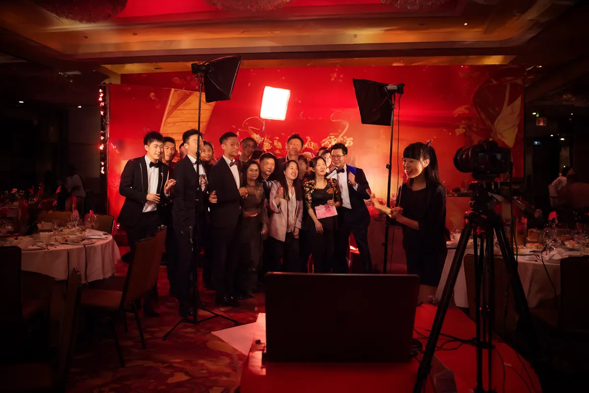 公司年会现场的红金色开放式拍照亭，工作人员正在引导同事们合影